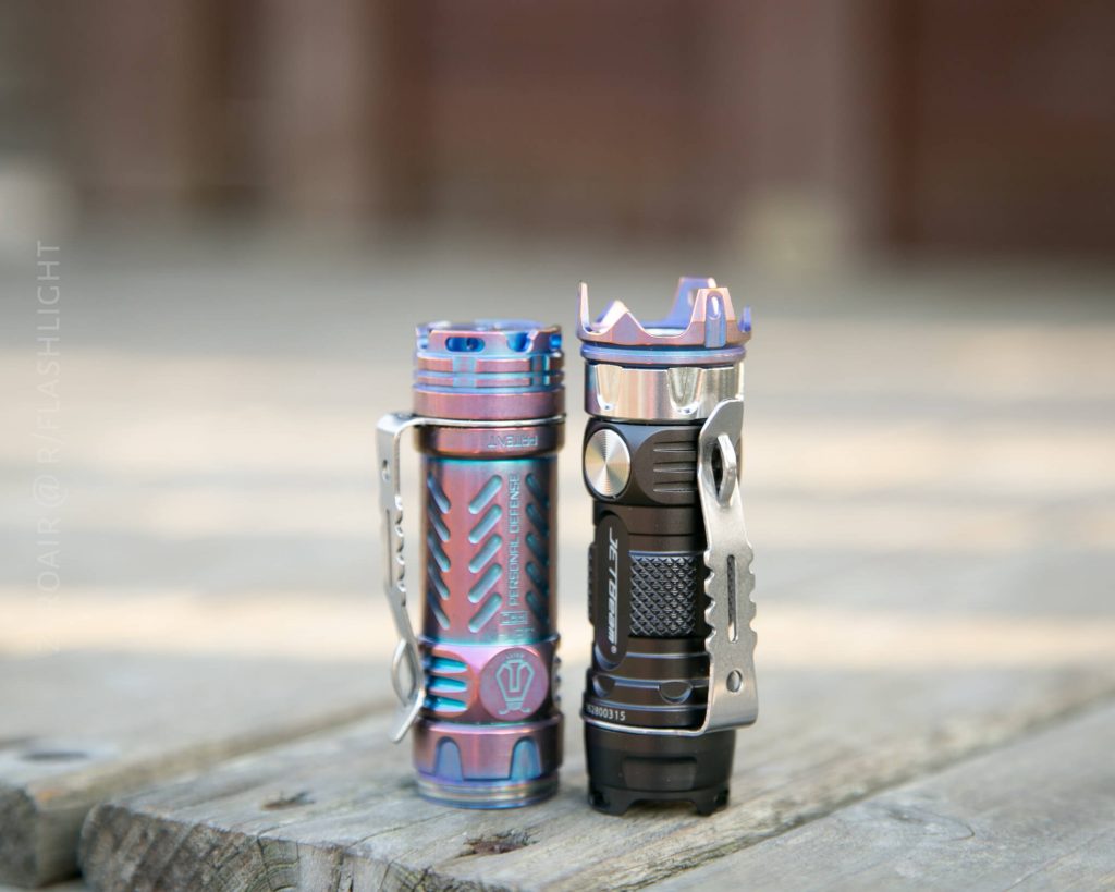 Two compact, metal flashlights with grooved designs stand upright on a wooden surface. One flashlight has a blue and purple iridescent finish, while the other is dark grey with silver accents.