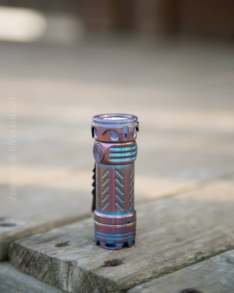 A metallic flashlight with a multicolored, iridescent finish stands upright on a wooden surface outdoors. The design includes grooves and textured patterns on its body.