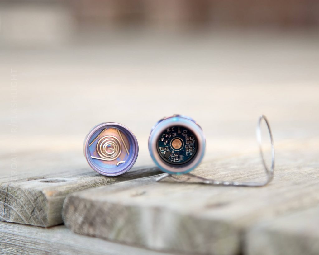 A close-up of a disassembled flashlight shows its head and tail cap placed on a wooden surface, revealing the metal spring and electronic components inside each part.