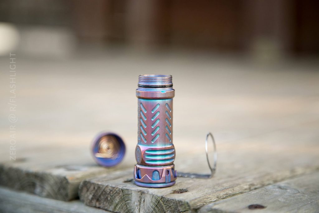 A metallic, cylindrical flashlight with a textured grip is shown standing upright on a wooden surface. Its tailcap is unscrewed and placed beside it, exposing the battery compartment and spring.