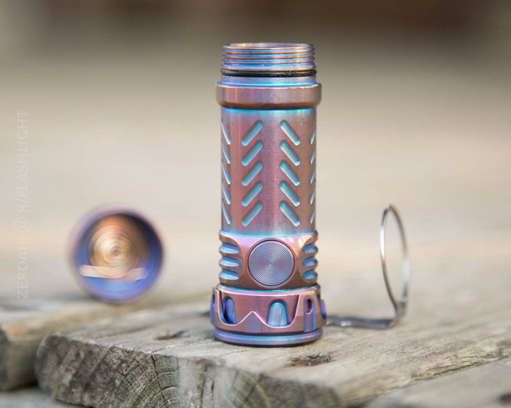 A metallic, cylindrical flashlight body with a threaded top and a textured grip sits upright on a wooden surface. The flashlight’s tail cap is detached and lying nearby, showing the battery compartment.