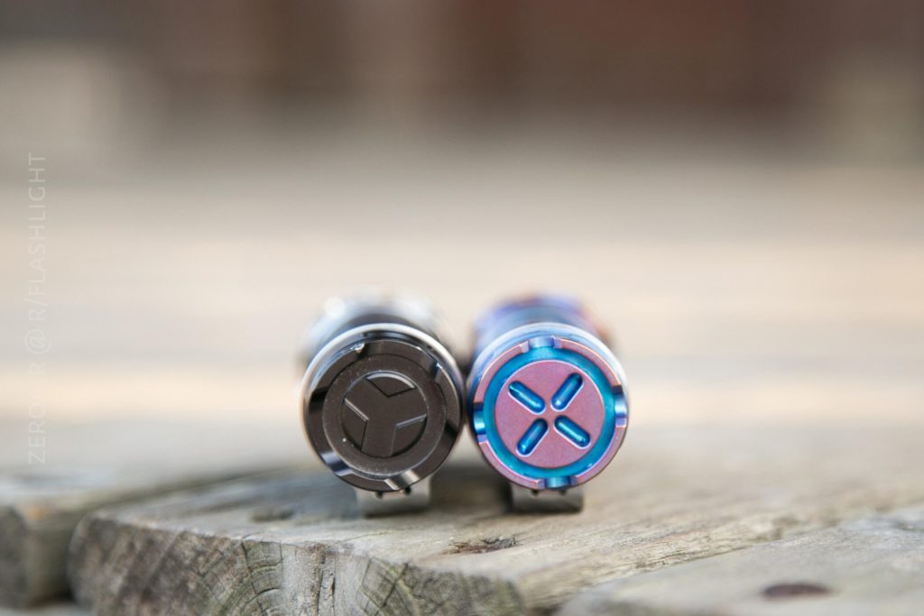 Two small cylindrical flashlights lie side by side on a wooden surface, with their tail ends facing the camera. The left one is dark gray, and the right one is blue and purple with metallic finishes.