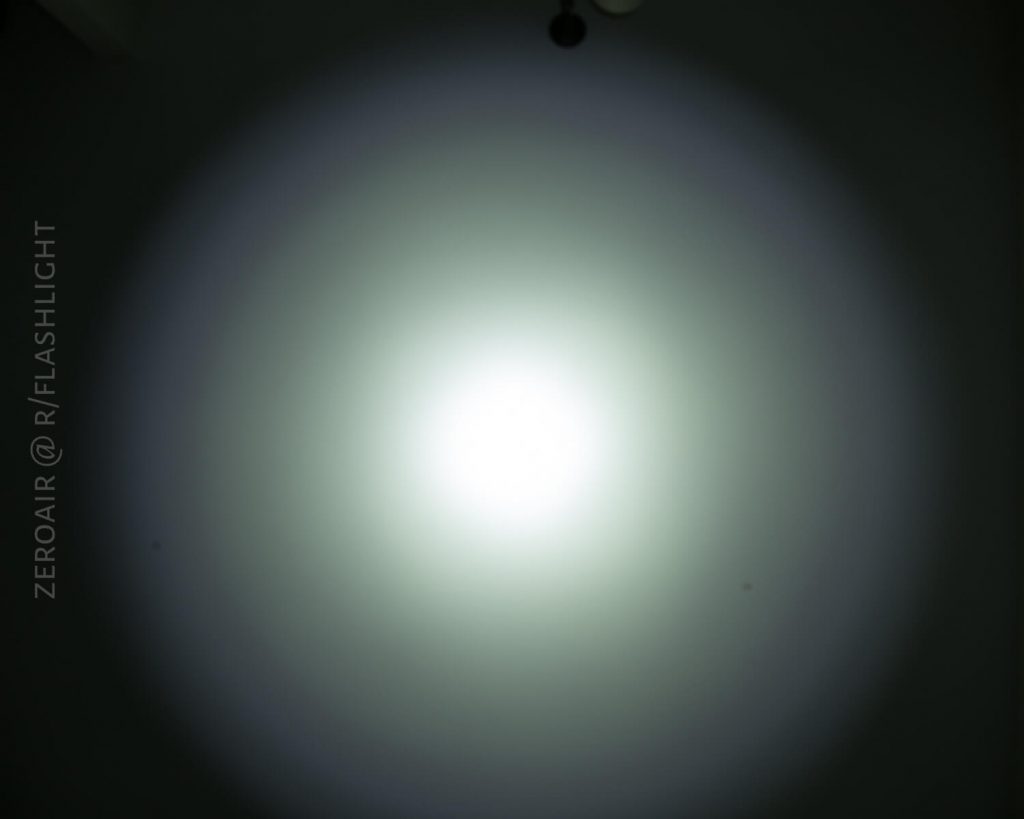 A bright white flashlight beam creates a circular spot on a dark surface. The center is intensely bright, fading gradually to dimmer light at the edges. Text along the left side reads “ZEROAIR @ R/FLASHLIGHT.”