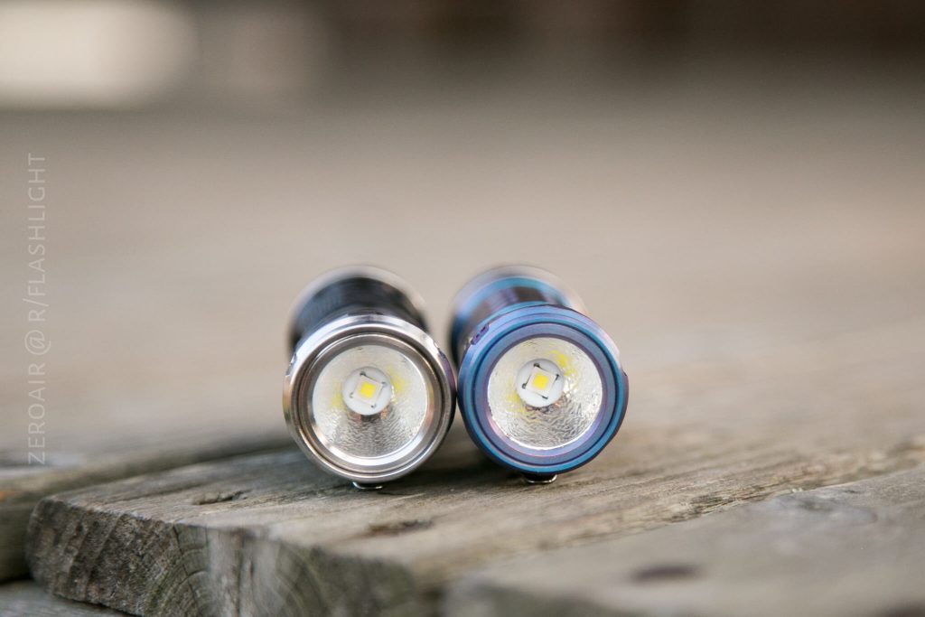 Two LED flashlights are placed side by side facing the camera on a wooden surface. The flashlight on the right has a blue accent around the lens, while the one on the left has a silver accent.