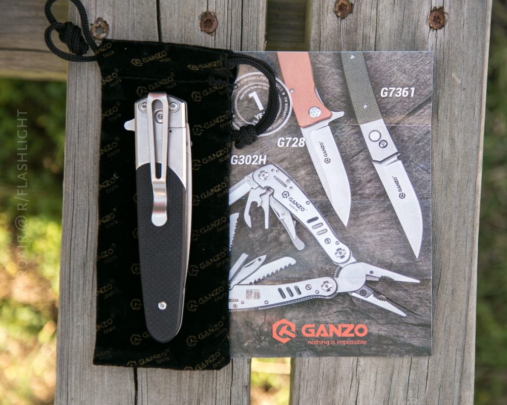 A folded Ganzo pocket knife with a black handle rests on a branded black pouch, next to a Ganzo product brochure displaying various knives and multitools on a wooden surface.