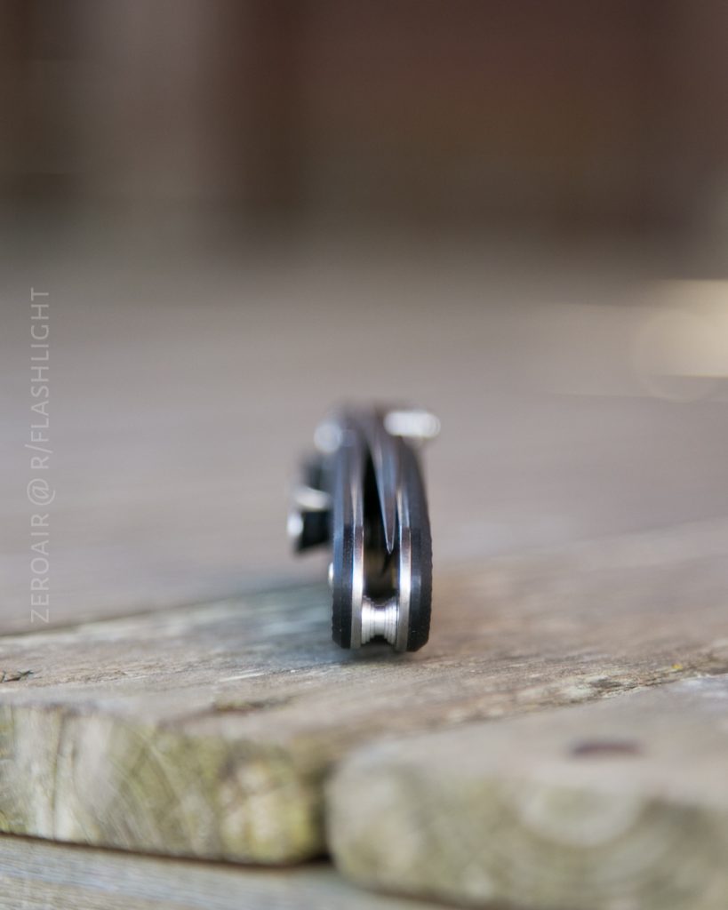 A folding pocket knife is shown closed and standing on its edge on a wooden surface. The background is blurred, and the knifes handle and blade spine are in focus. Text reads, ZEROAIR @ FLASHLIGHT.