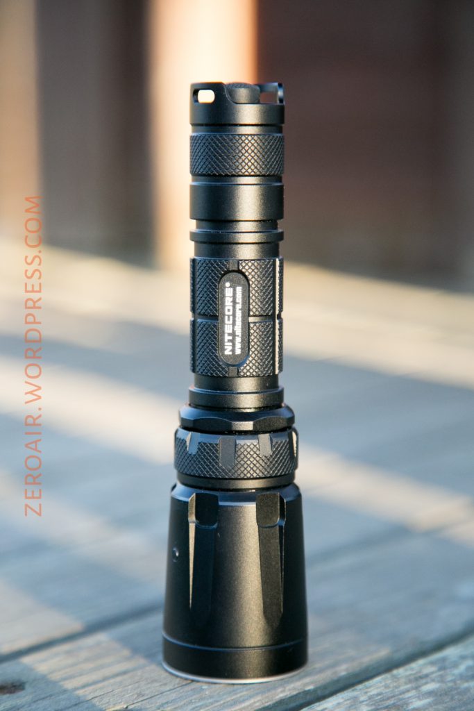 A black Nitecore flashlight is standing upright on a wooden surface, photographed in natural light. The brand label is visible on the flashlight’s body. The background is softly blurred.