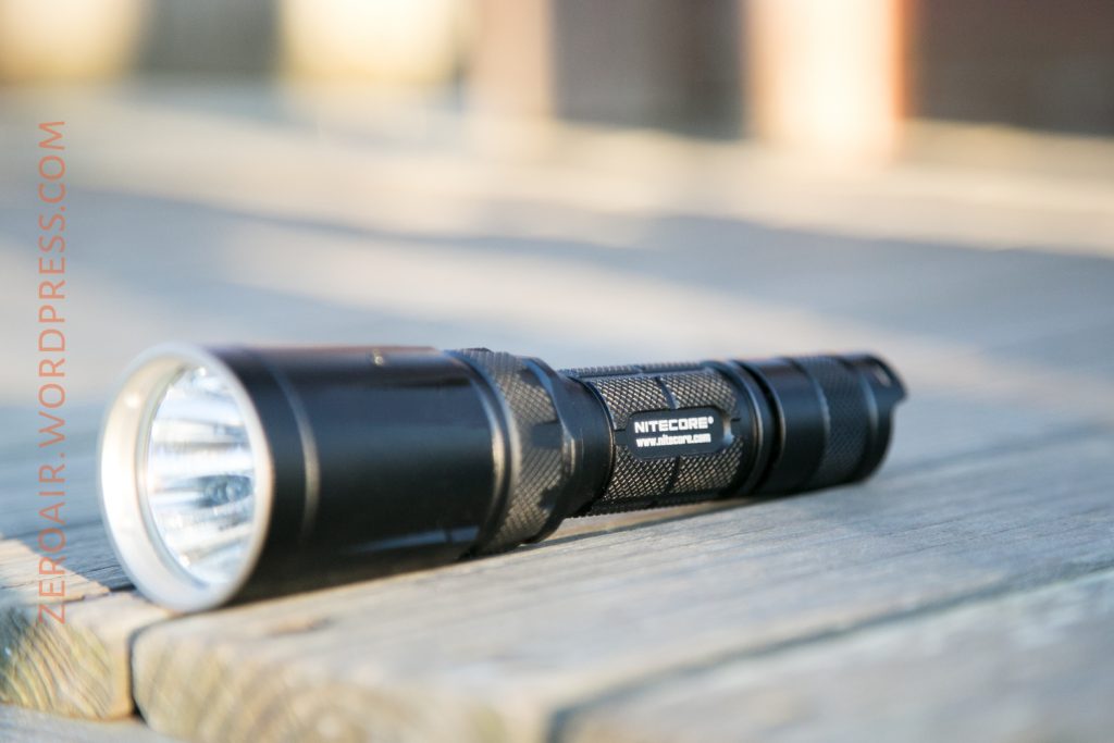 A black Nitecore flashlight is lying on a wooden surface outdoors. The flashlight is in focus, positioned at an angle, with blurred background and sunlight casting soft shadows.