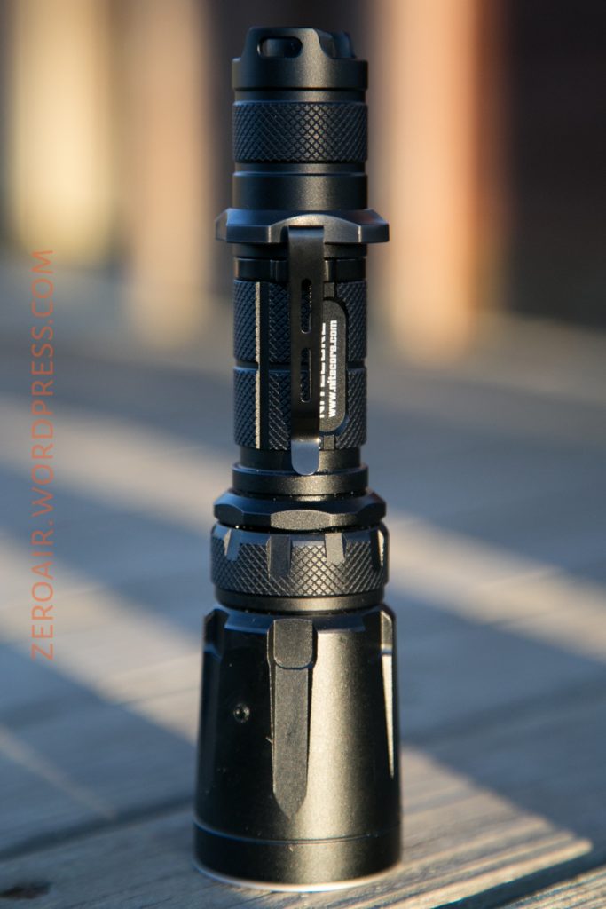 A black tactical flashlight stands upright on a wooden surface, with metal ridges for grip and a side clip. The background is out of focus, and the text ZEROAIR.WORDPRESS.COM is visible along the left side.