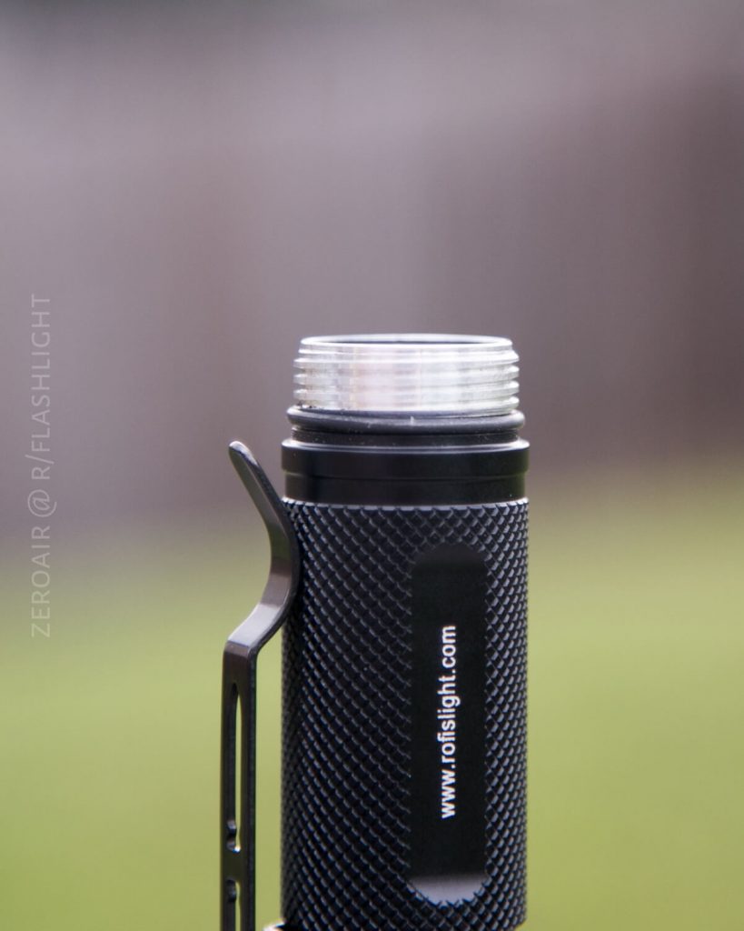 A close-up of a black textured flashlight with its head removed, showing metal threads at the top. The flashlight has a clip and a website URL printed on its side. The background is blurred.