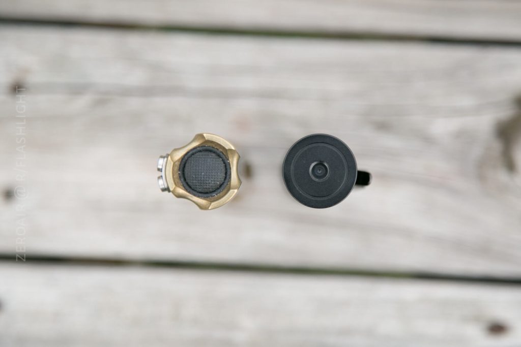 Top-down view of two flashlight tail switches on a wooden surface; the left switch is gold with a textured black button, while the right switch is black and smooth.