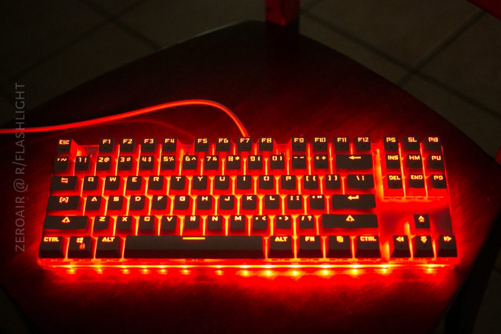 A mechanical keyboard with red backlighting sits on a dark wooden surface. The keyboard is illuminated, and its keys are clearly visible. A faint watermark appears on the left side of the image.