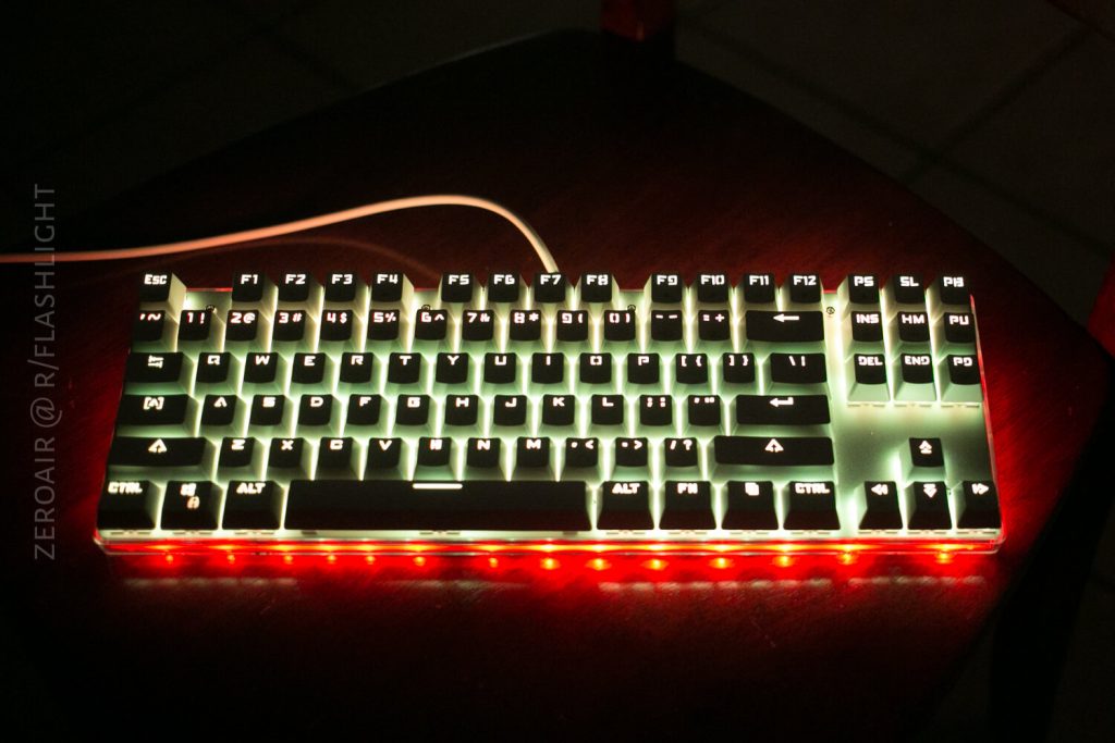 A white mechanical keyboard with illuminated keys sits on a dark wooden surface. The keyboard glows with white backlighting on the keys and red lighting around the base. A white cable extends from the back.
