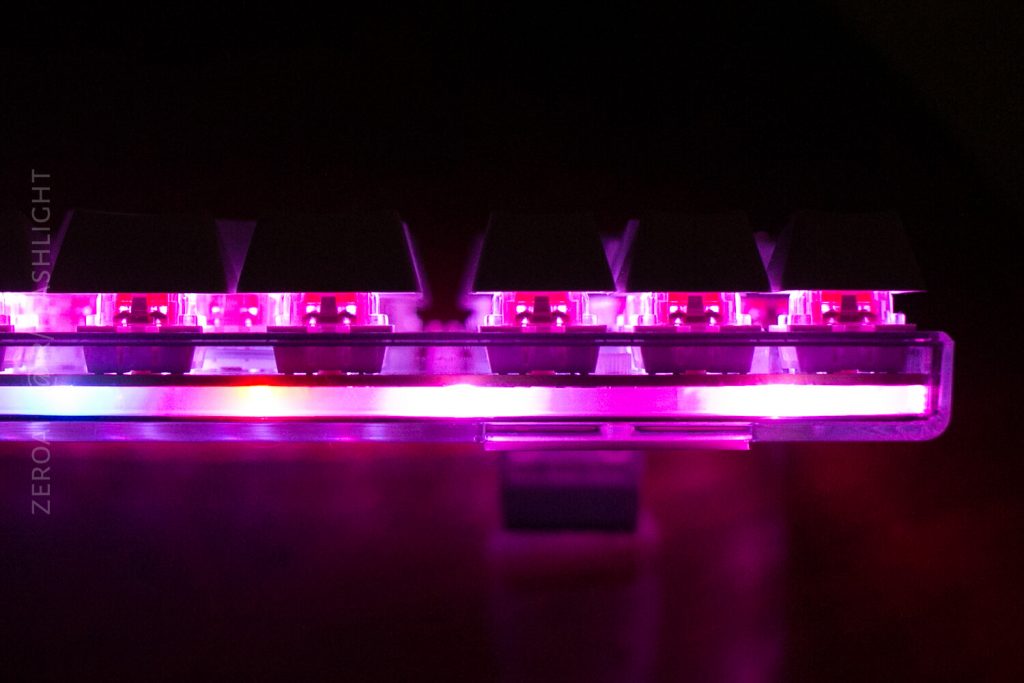A close-up side view of a mechanical keyboard with pink and purple backlighting illuminating the clear switch housings and base, set against a dark background.