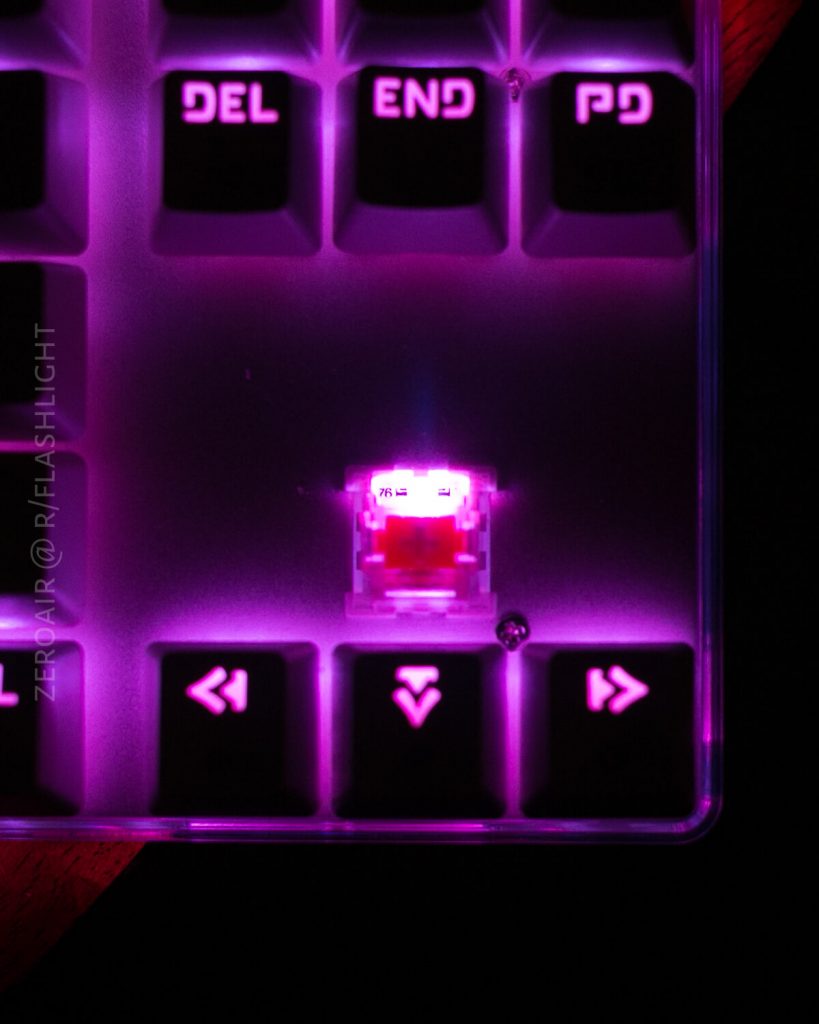 A close-up of a backlit mechanical keyboard with magenta lighting shows a removed keycap, revealing a red switch. The surrounding keys are illuminated and labeled “DEL,” “END,” PD, and arrow keys.