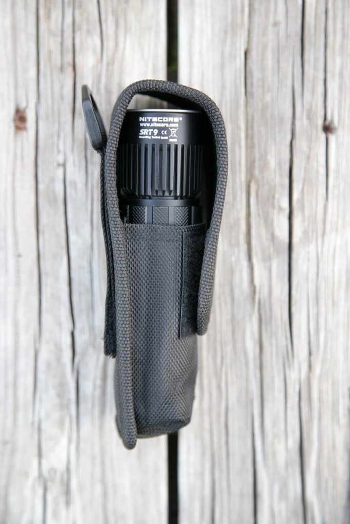 A black Nitecore flashlight is partially visible inside a black textured nylon holster, placed vertically on a weathered wooden surface.