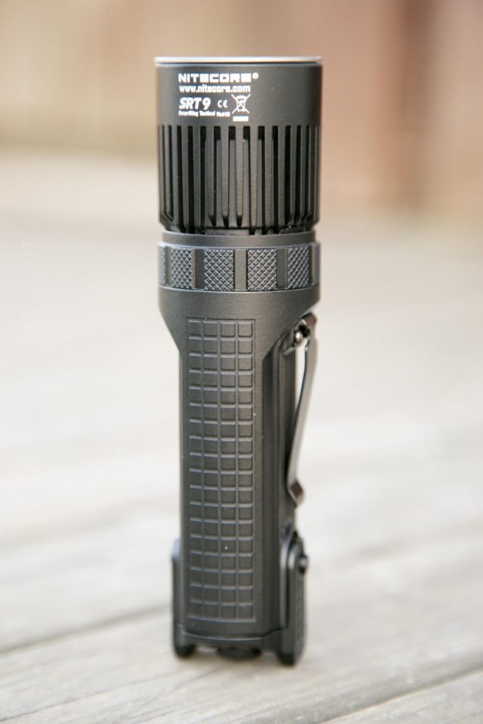A black Nitecore SRT9 flashlight stands upright on a wooden surface, displaying its textured grip, side pocket clip, and branding on the top section. The background is blurred.