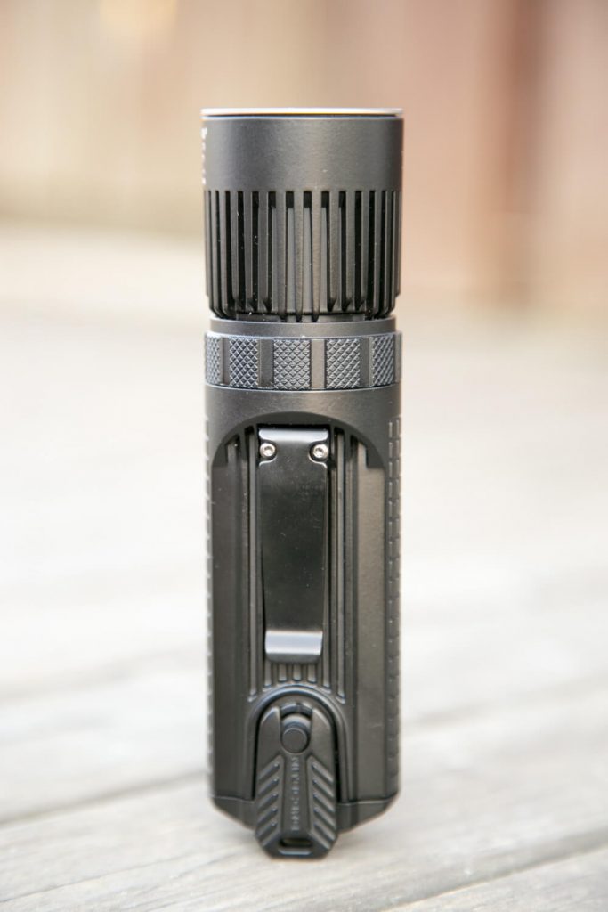 A black LED flashlight standing upright on a wooden surface, showing its side with a metal pocket clip and textured grip.