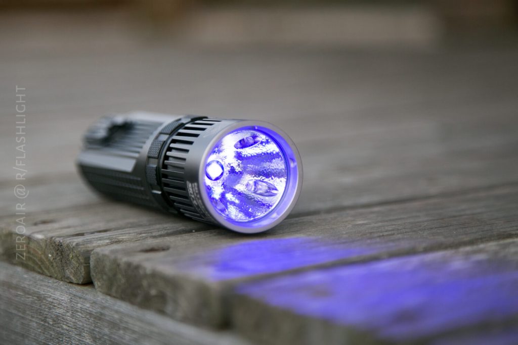 A black flashlight lies on its side on a wooden surface, emitting a bright blue-violet light that illuminates the wood in front of it.