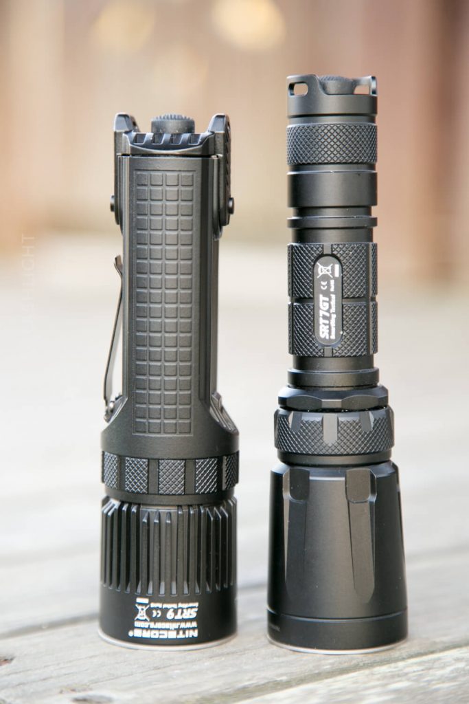 Two black tactical flashlights standing upright on a light-colored surface. The flashlight on the left is shorter with a textured grip, while the one on the right is taller with a larger head and labeled SRT7.