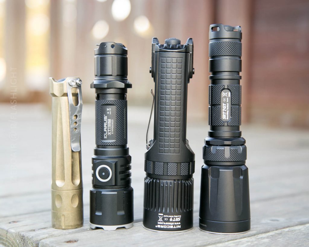 Four flashlights of varying sizes and designs, made from metal, stand upright in a row on a wooden surface with a blurred brown background. Each flashlight features unique grips, buttons, and ridges.