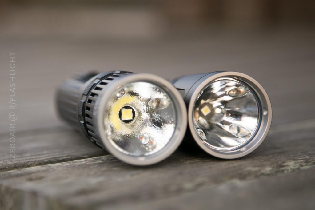 Two flashlights lie side by side on a wooden surface, with their lenses facing the camera. The left reflector has a textured surface, while the right one is smooth with visible LED components.
