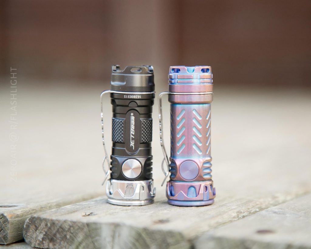 Two compact flashlights stand upright on a wooden surface. The left flashlight is black and silver with a JETBeam label, while the right one has a metallic, purple-blue finish. Both have metal clips attached.