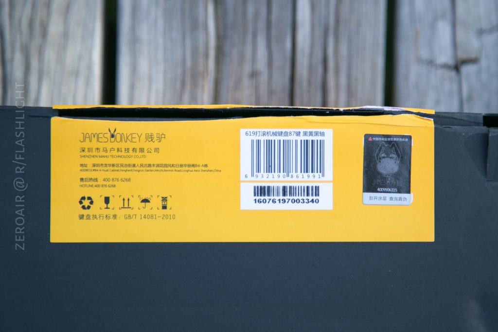 A close-up of a black and yellow product box with text in both Chinese and English, a barcode, multiple symbols, and a small QR code. The box is placed on a wooden surface.