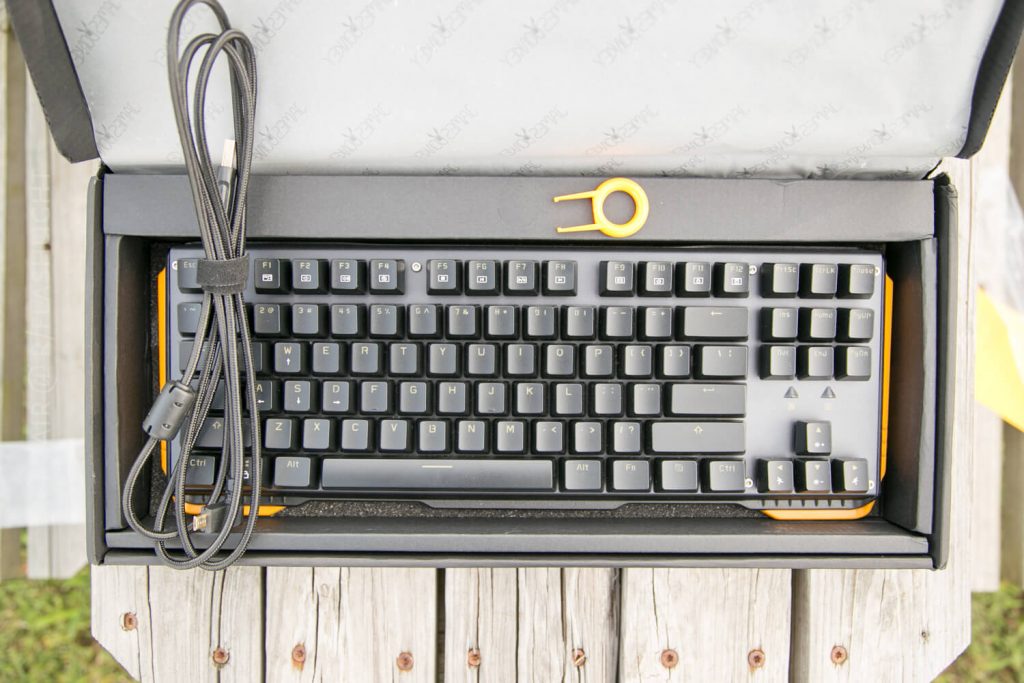 A mechanical keyboard in a box with a braided USB cable and a yellow keycap puller, displayed on a wooden surface. The keyboard has black keys and orange trim.