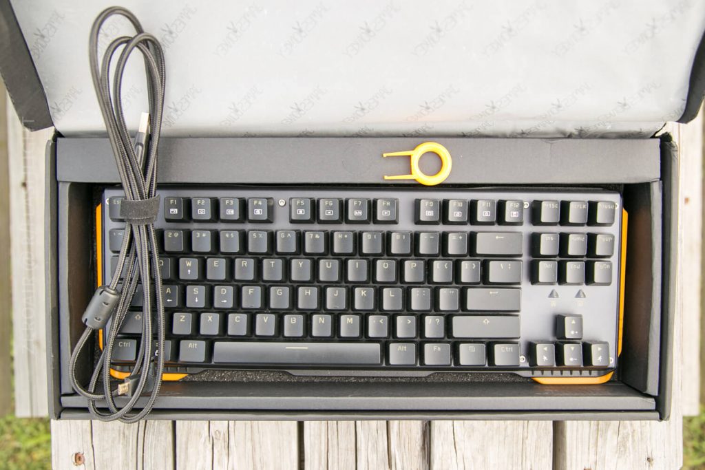 A black mechanical keyboard in a box with a detachable braided USB cable and a yellow keycap puller placed on top of the keyboard, resting on a wooden surface.