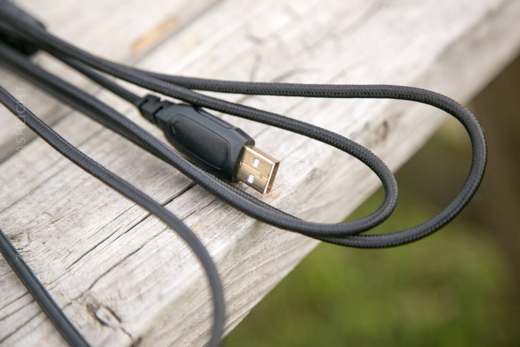 A close-up of a black USB-A cable lying on a weathered wooden surface, with greenery blurred in the background.
