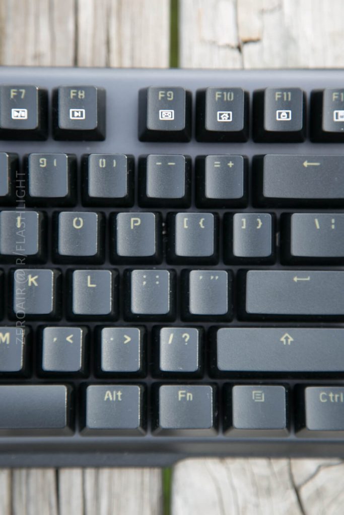 Close-up of a black computer keyboard showing the F7 to F12 keys on the top row and the letter keys from I to L, with visible wear on the Alt, Fn, and right arrow keys. The keyboard is on a wooden surface.