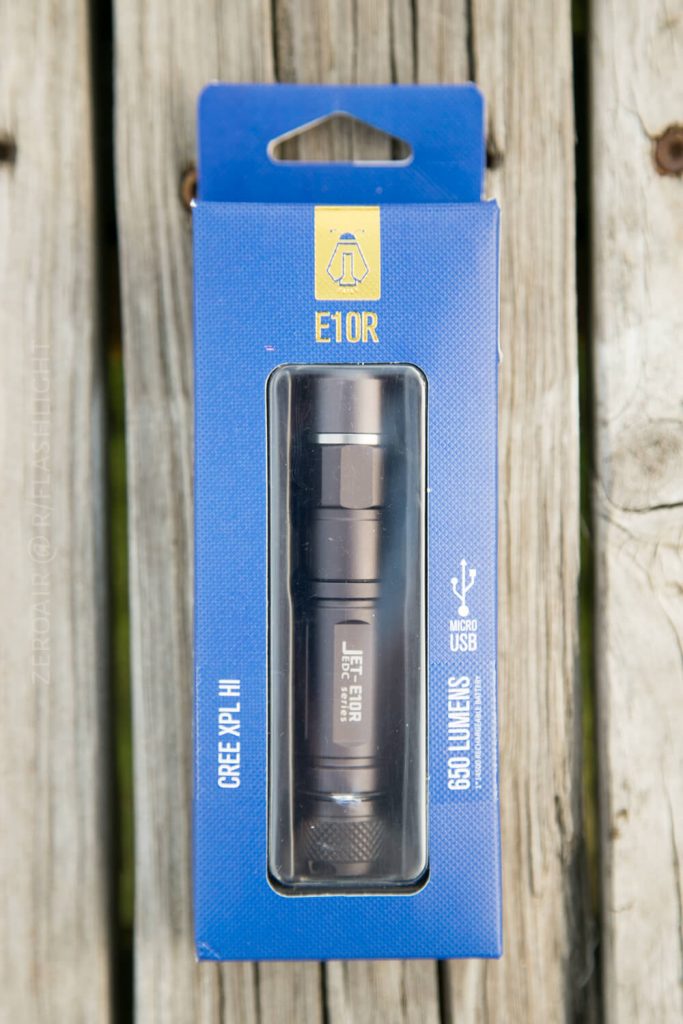 A black flashlight is displayed in blue packaging labeled E10R with icons for micro USB charging and LED light. The box is set on a wooden surface.