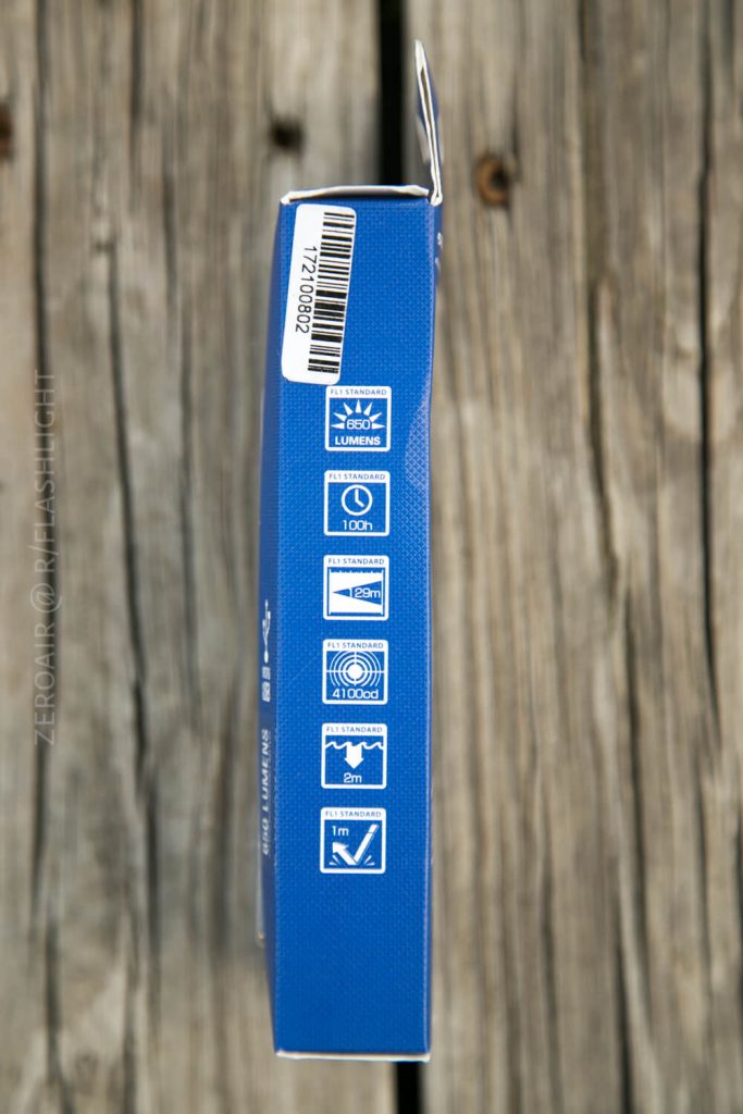 A blue product box stands upright on a wooden surface, displaying icons indicating 100 lumens brightness, 4,100 candela, 100 hours runtime, a 5-meter waterproof rating, and a 1-meter drop resistance. A barcode is visible on top.