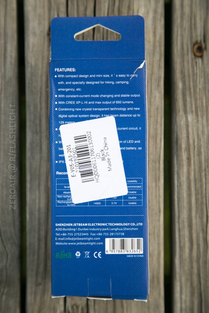 A blue product box stands upright on a wooden surface. Its back side lists product features, a barcode label, and manufacturing details, including Made in China and Shenzhen Jetbeam Electronic Technology Co., Ltd.