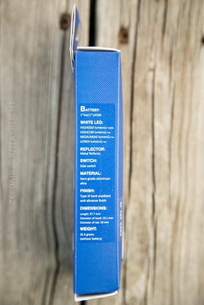 The side of a blue product box lists specifications such as battery type, white LED details, reflector, switch, material, finish, weight, and dimensions. The box is on a wooden surface.