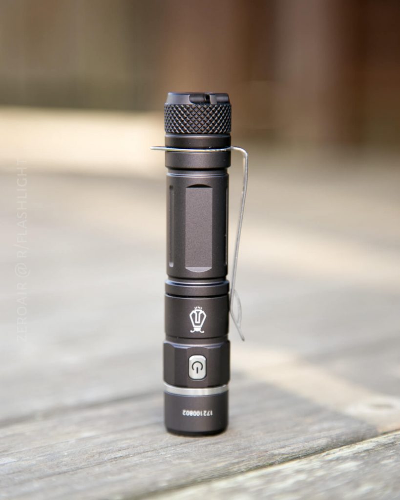 A black compact flashlight with a textured grip and pocket clip stands upright on a wooden surface. The flashlight has a side button and visible branding with serial numbers on its body.