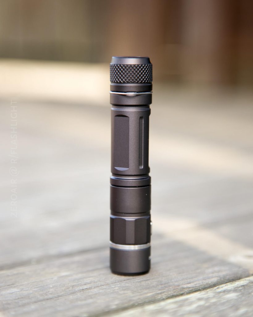 A compact black flashlight stands upright on a wooden surface, with a blurred background. The flashlight has a textured grip near the top and metallic rings near both ends.