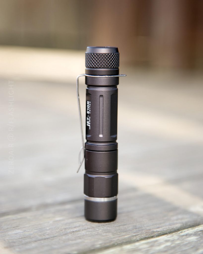A compact black flashlight with a metal pocket clip stands upright on a wooden surface. The background is blurred, drawing focus to the flashlight.