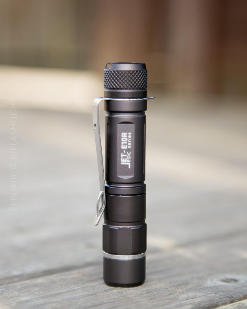 A small, black, cylindrical flashlight with a metal pocket clip stands upright on a wooden surface. The flashlight is labeled “JET-E21A” on the side. The background is out of focus.