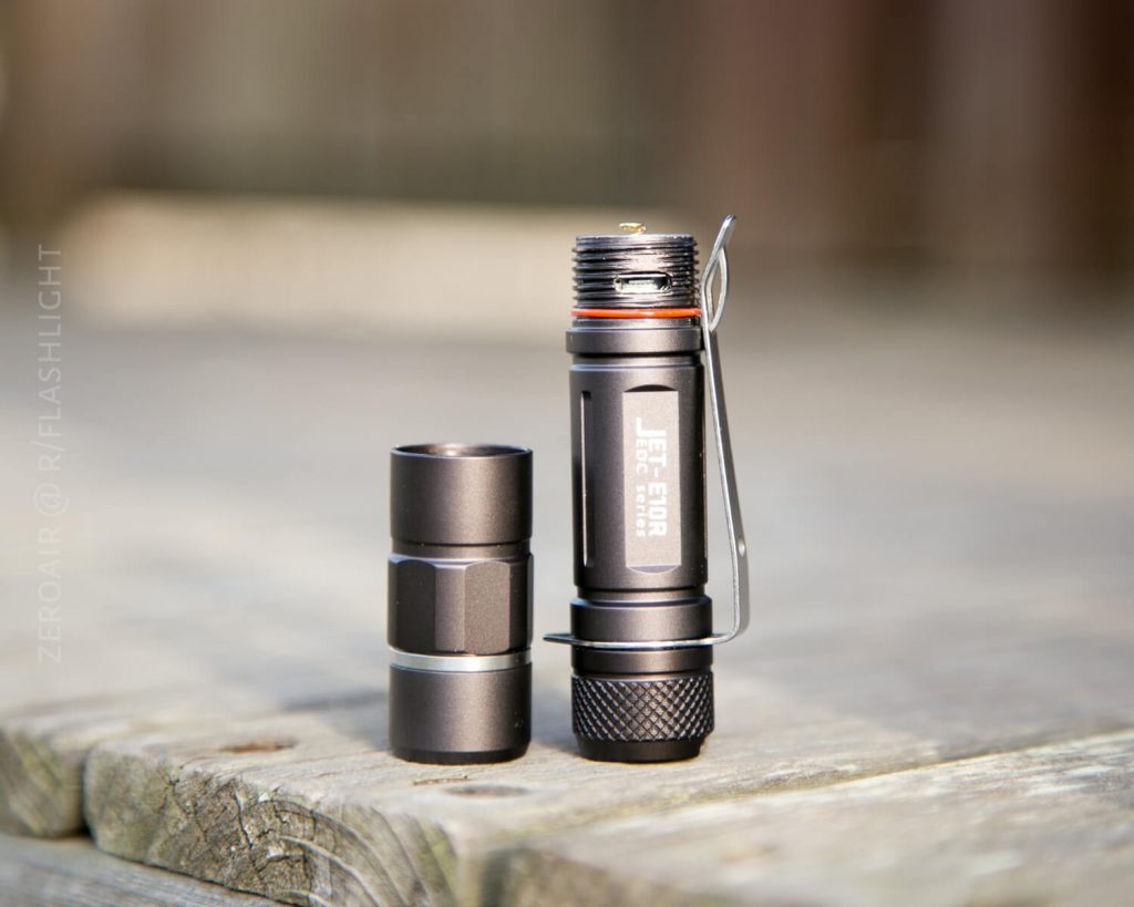 A small black Jet-Eater flashlight stands upright on a wooden surface with its battery compartment unscrewed and placed beside it. The photo is bright and in focus, with background out of focus.