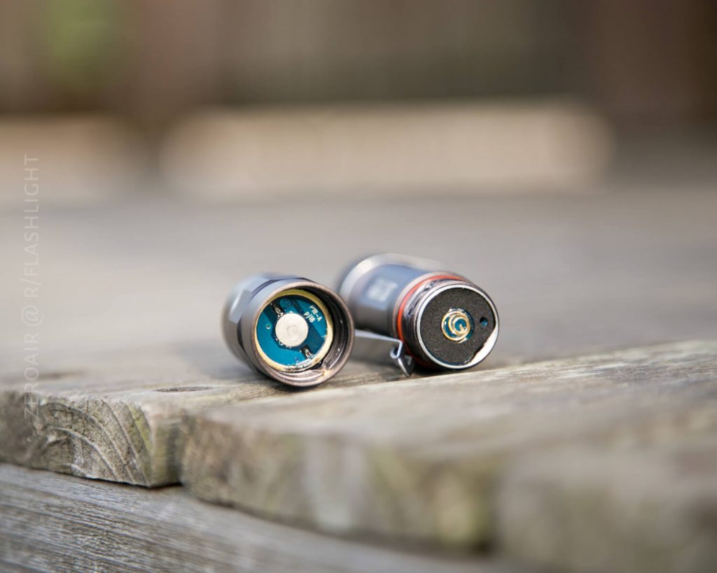 Two cylindrical batteries lie on a wooden surface, both showing their metal terminals. One battery displays its positive end, while the other shows its exposed circuit board. The background is blurred.
