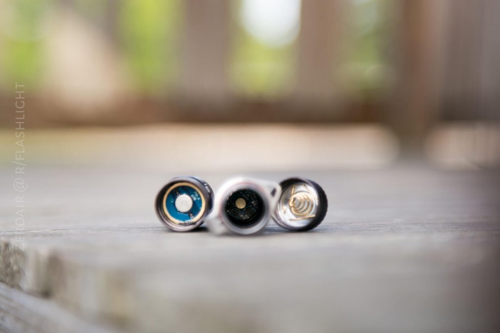 Three flashlight bodies are shown lying horizontally on a wooden surface, with the battery compartments facing the camera and their contacts clearly visible. The background is blurred.