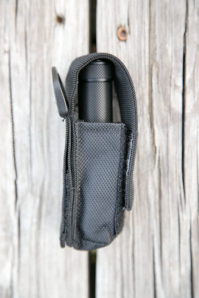 A black flashlight in a textured nylon holster with a flap, photographed from above on a weathered wooden surface.