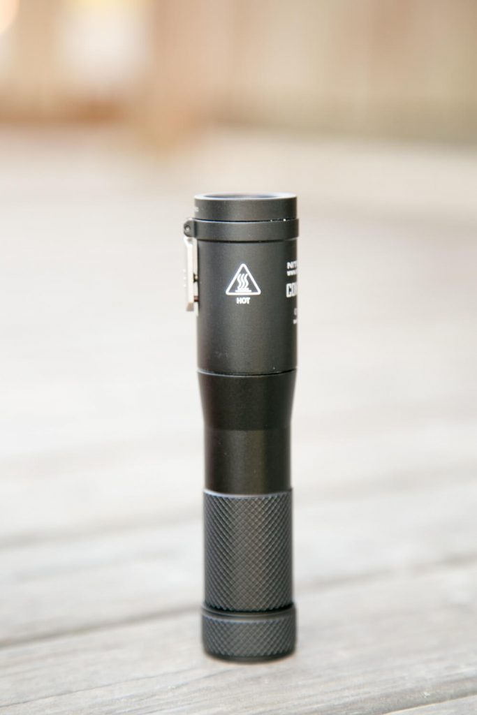 A black flashlight standing upright on a wooden surface. The flashlight has textured grip detailing on the lower part and a warning symbol indicating heat on the upper part. The background is out of focus.