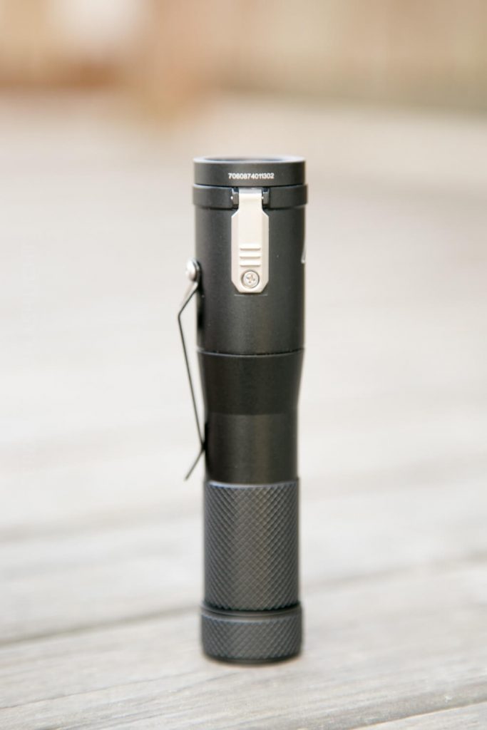 A black handheld flashlight with a textured grip and belt clip is standing upright on a wooden surface. The flashlight has a visible serial number on the top rim.