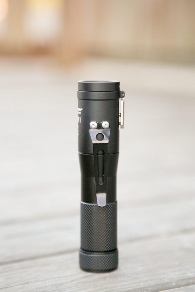 A black, compact flashlight with a textured grip and metal clip stands upright on a wooden surface. The background is blurred in neutral tones.