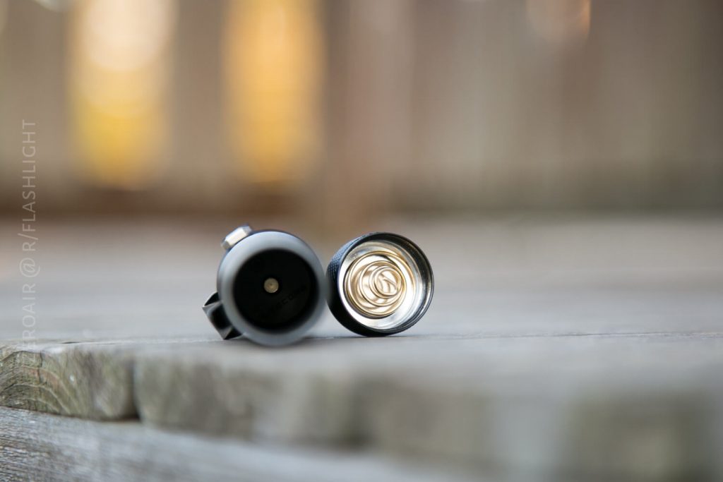 A close-up of a disassembled flashlight lying on a wooden surface, showing the inside with a visible metal coil spring in the battery compartment. The background is blurred.
