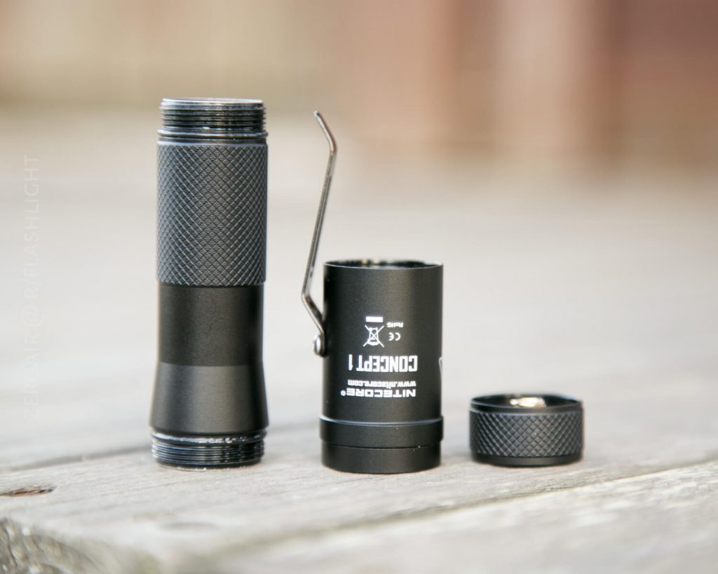 A disassembled black flashlight with three parts—the main body, LED housing with a metal clip, and a screw-cap—are arranged on a wooden surface.