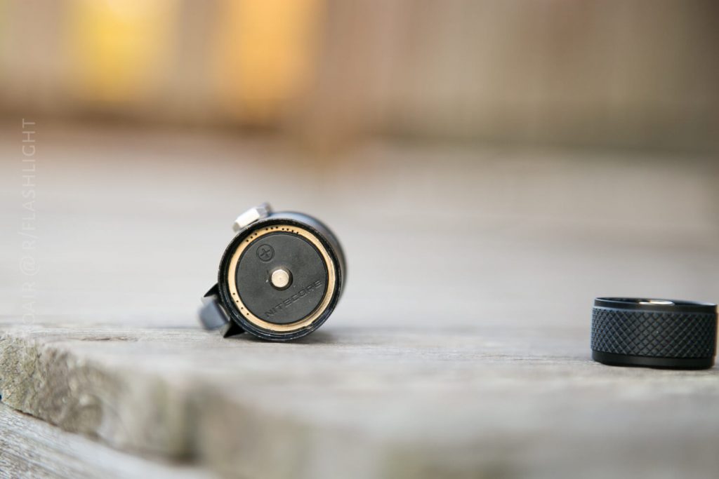 A close-up of a black flashlight lying on its side on a wooden surface, with its battery compartment open and the end cap placed nearby. The photo has a blurred background.